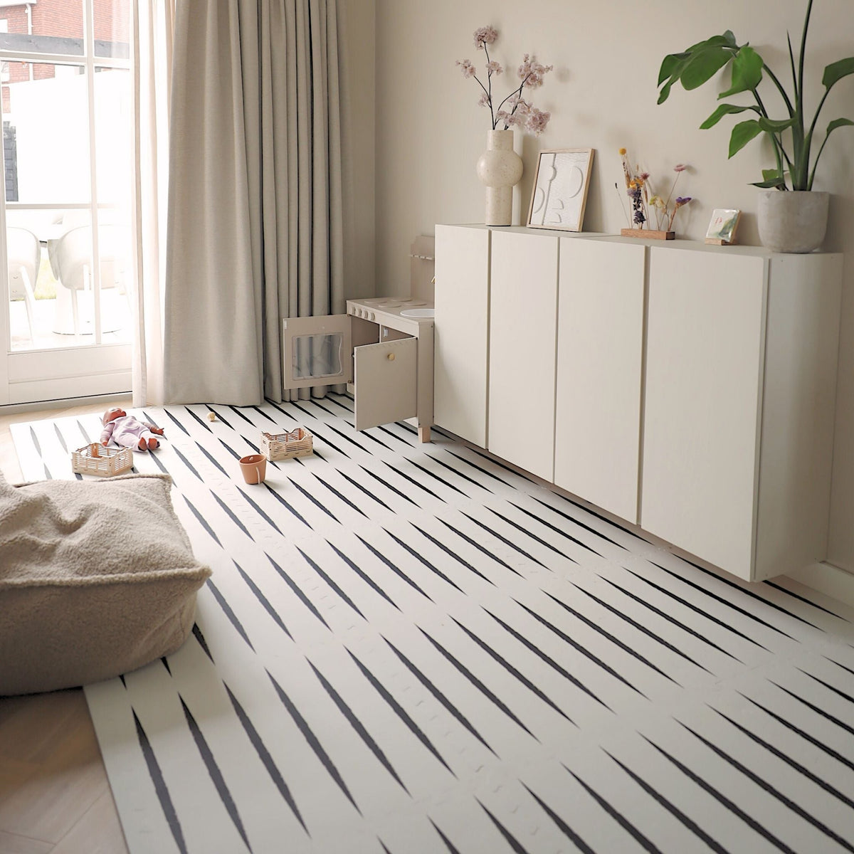Tapis de jeu Lucio Ecru posé sur un parquet clair, dans un salon avec un pouf et des rangements